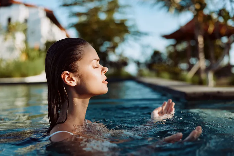 Young woman with wet hair relaxing in outdoor pool with closed eyes enjoying sunlight and water. Concept of wellness, lifestyle, and summer leisure in natural environment.