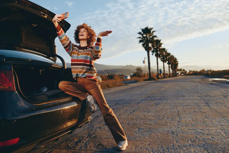 Woman lifestyle by car outdoors at sunset, curly hair and striped casual outfit, jumping pose on cracked road, palm trees lining the street, warm golden light, carefree