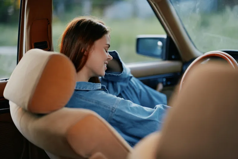 A young woman in casual denim clothing enjoys a relaxed moment in a vintage car, embodying a carefree summer vibe with a serene expression.
