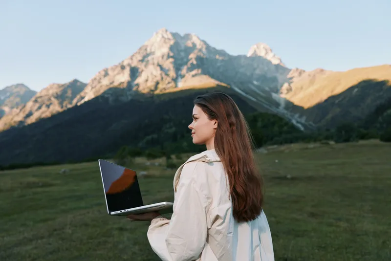 A woman stands outdoors with a laptop in hand, overlooking a scenic mountain landscape. She wears light outerwear and looks focused yet relaxed, blending technology with nature in a calm, productive
