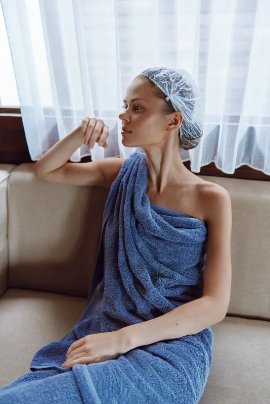 Relaxed woman wrapped in blue towel and wearing shower cap sitting on sofa near curtain, natural light highlighting calm and refreshed skin in cozy indoor setting.