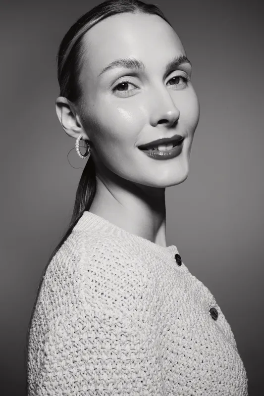 Elegant caucasian woman with fair skin and dark hair smiles softly wearing knitted sweater and hoop earrings in studio, soft lighting highlights facial features.