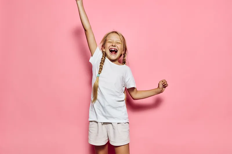 Happy girl with braided hair wearing white clothes smiling and cheering on a pink background. Studio shot with bright lighting.