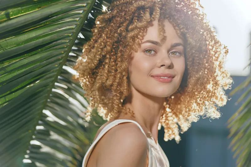 smiling woman with curly hair enjoys sunny outdoor setting between green palm leaves, showcasing natural beauty and warm summer vibes with soft sunlight.