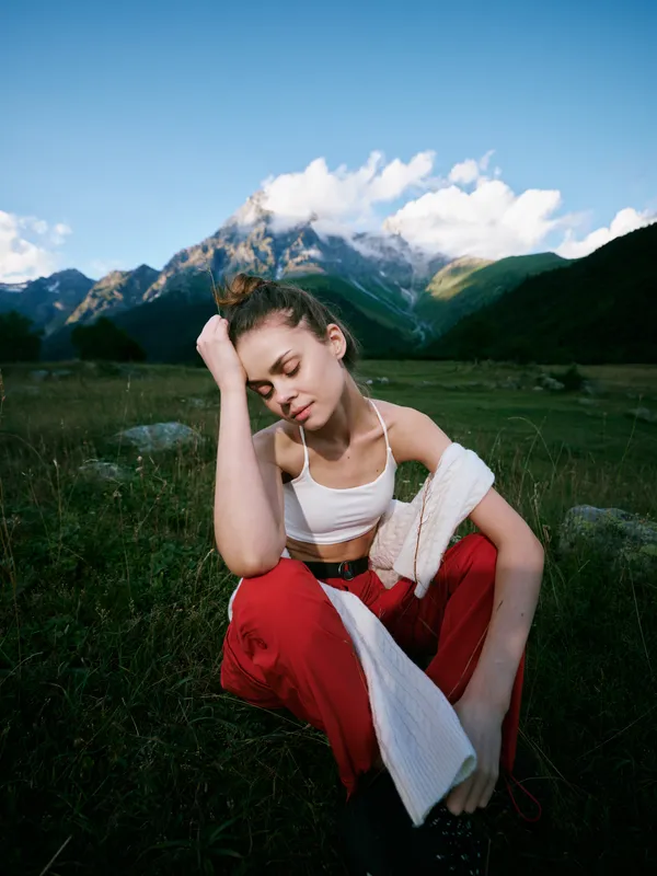 Outdoor scene shows a tired woman resting on a grassy field with mountains and blue sky, wearing a white crop top and red pants, towel draped over her shoulder