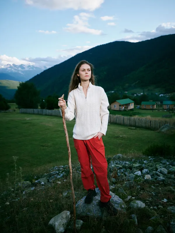 Woman standing on a rocky overlook with a wooden staff, green valley and distant mountains behind, casual white top and red pants, capturing an adventurous yet peaceful moment in nature