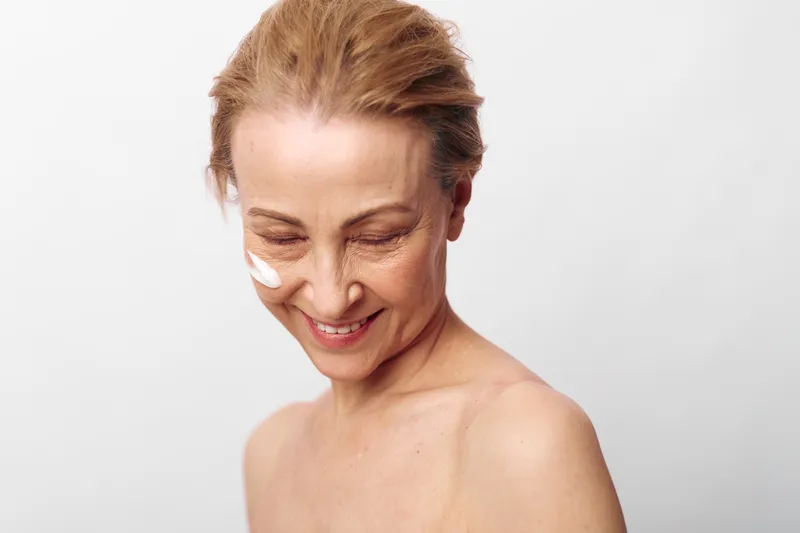 Smiling mature woman with natural skin and cream on cheek looks down on white background. Concept of skincare, aging beauty, self-care, and natural wellness for older adults.