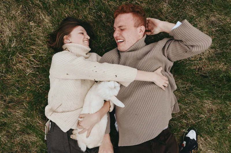 Couple lifestyle goat kid outdoors grass sweater cozy affection nature, a candid moment as they lie together in the sun, cradling a white kid while smiles rise in a peaceful rural setting.