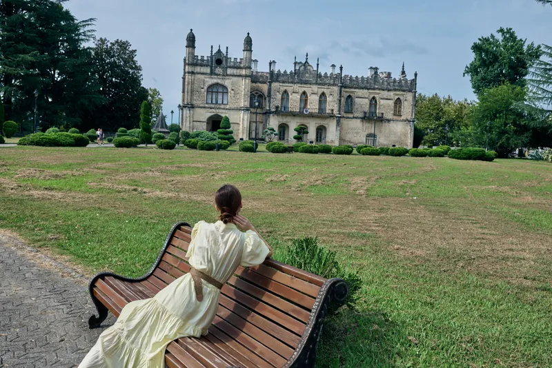 woman in cream vintage dress sits on a curved wooden bench in a manicured park with a historic castle in the background, conveying a serene and timeless countryside moment