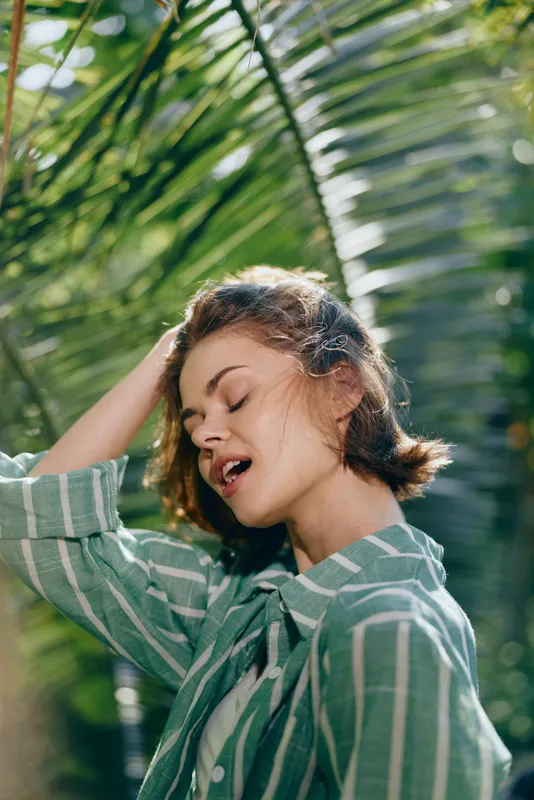 A young woman enjoys warm sunlight outdoors, eyes closed, hair lifted by breeze, posing in a lush tropical setting.