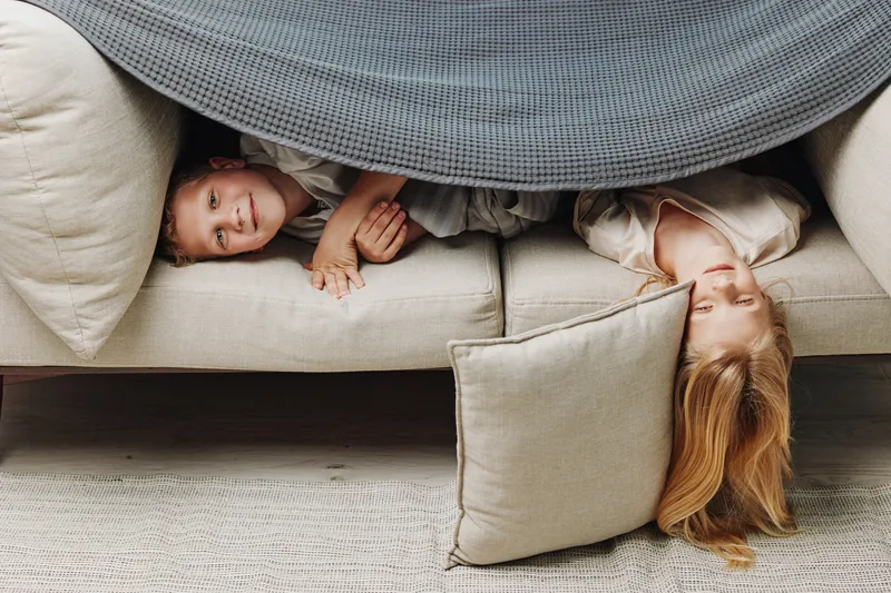 Two children lying under grey blanket on beige couch, smiling and playing indoors. Cozy home environment with soft cushions and warm carpet floor.