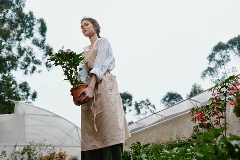 Young woman holding a potted plant in a greenhouse, wearing a casual outfit and an apron, with a serene expression reflecting her passion for gardening