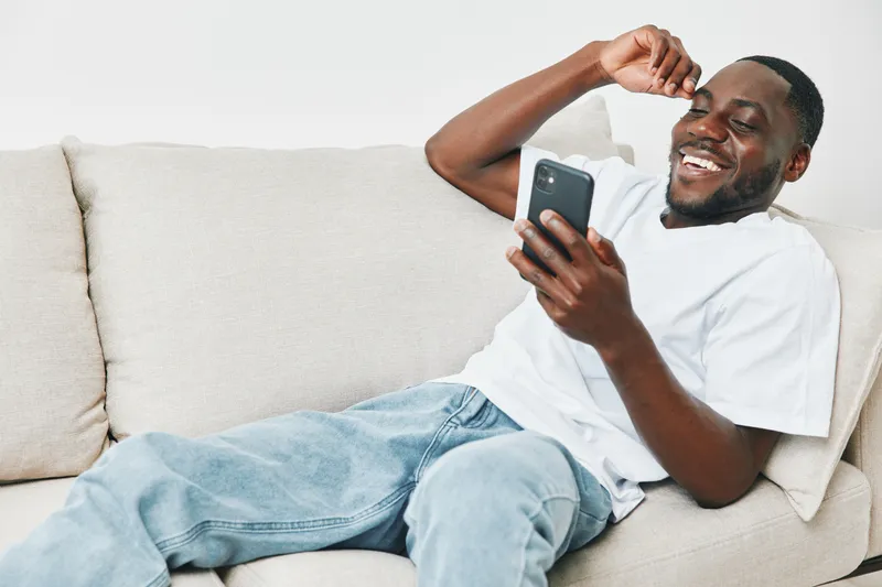 Happy man enjoying smartphone on a light couch, wearing casual white t shirt and jeans, conveying joy and relaxation in a modern home interior
