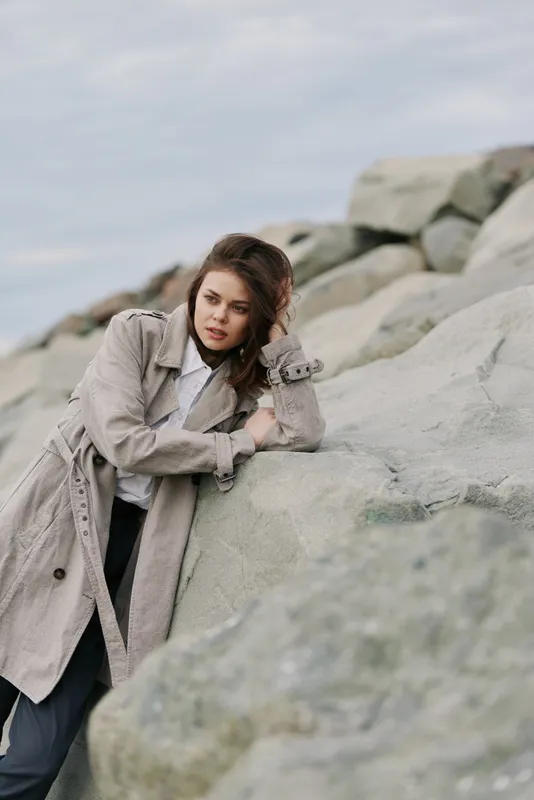 Young woman in long coat leans on large rocks outdoors. She looks thoughtful with loose hair and casual style. Natural light and cloudy sky create moody atmosphere by the shore.