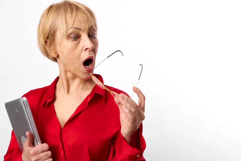 Woman elderly shocked expression holding tablet and glasses red shirt surprised look isolated in studio white background indoors soft light mouth open gaze to camera closeup portrait