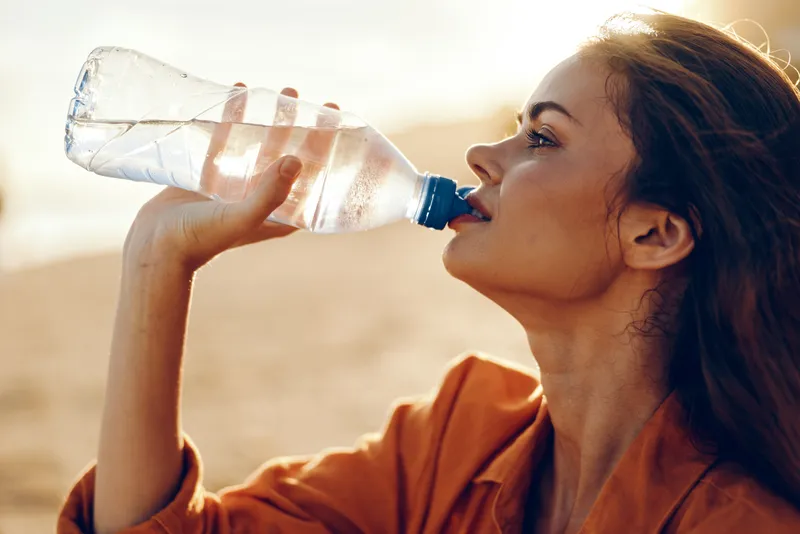 Woman drinking water outdoors in warm sunlight, side profile holding clear bottle, hydration and thirst relief, relaxed healthy lifestyle, soft breeze, sunlit ground, summer wellness