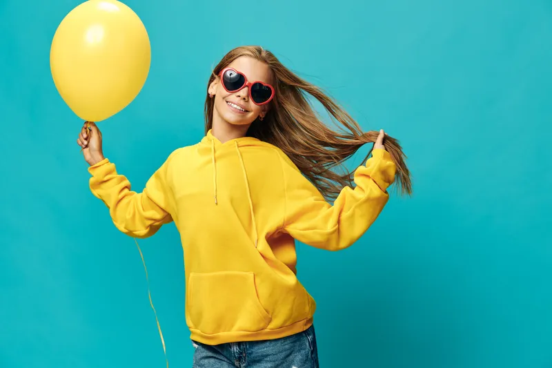 Young woman in yellow hoodie holding yellow balloon, wearing heart-shaped sunglasses, smiling and enjoying vibrant blue background with casual style and carefree mood.