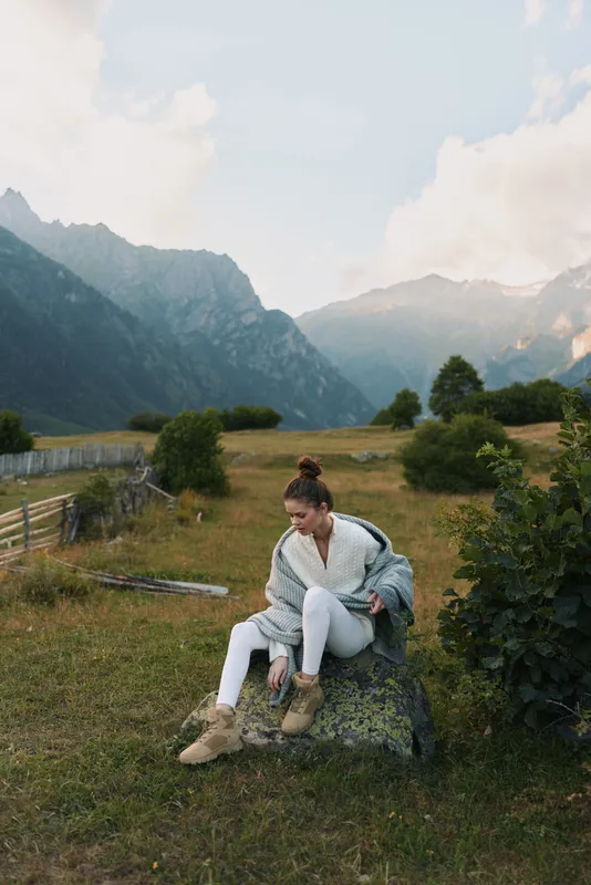 A young woman in white pants and beige boots sits on a rock in a meadow with distant mountains, a soft sweater visible, conveying calm solitude and contemplation.