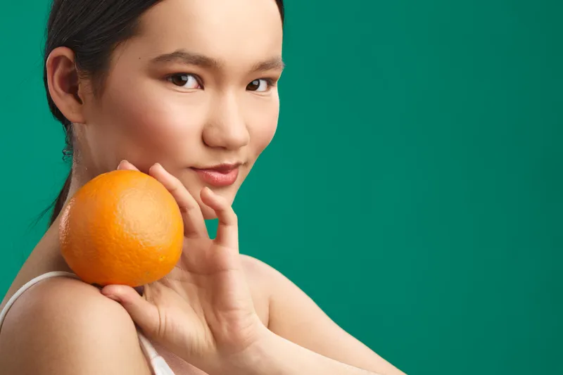 Asian woman holds an orange near her shoulder, smiling gently against a vivid green backdrop, conveying freshness, health, and a cheerful, confident mood.