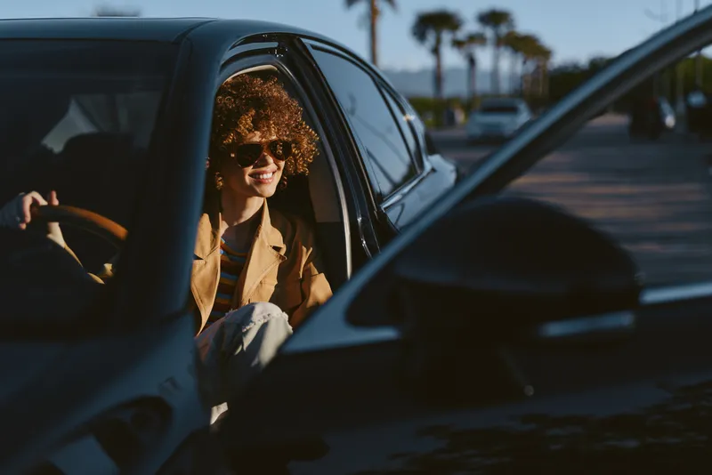 A woman sits in a car with the window down, enjoying sunlight and a retro film color vibe. A candid lifestyle moment conveying travel, nostalgia, and warm atmosphere.