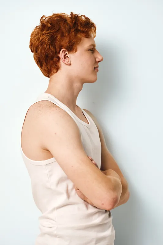 Young man with red curly hair wearing white tank top standing sideways with arms crossed against plain light background. Profile portrait of confident male with casual style.
