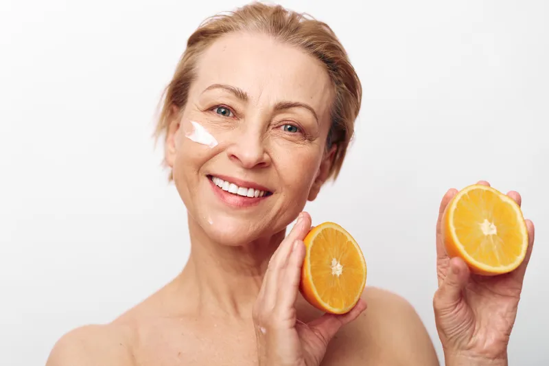 Smiling mature woman with natural skin holds fresh orange halves near face, applying white cream on cheek. Concept of healthy skincare, natural beauty, citrus fruit, and wellness treatment.