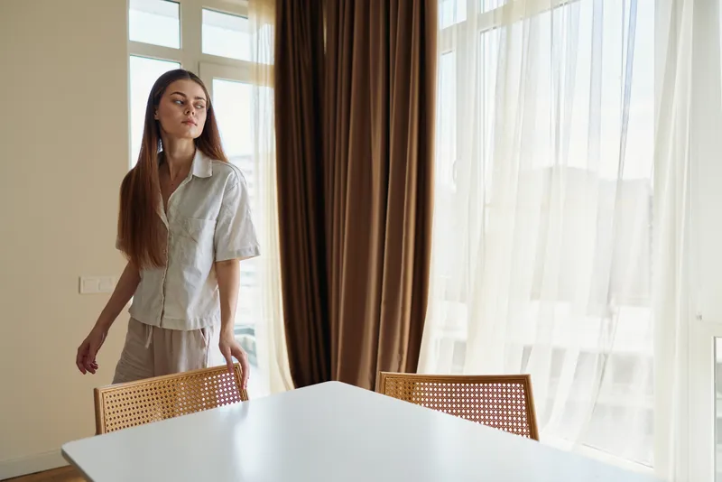 Woman standing by window with sheer curtains in bright home interior, looking thoughtful near dining table and rattan chairs, natural light creating calm domestic scene.