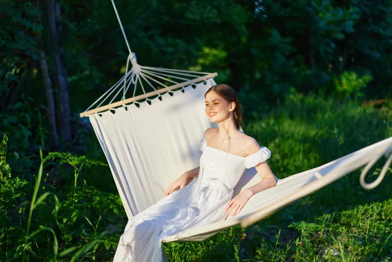 Young woman relaxing in a hammock in a lush green environment, wearing a white sundress, expressing tranquility and joy during a sunny summer day