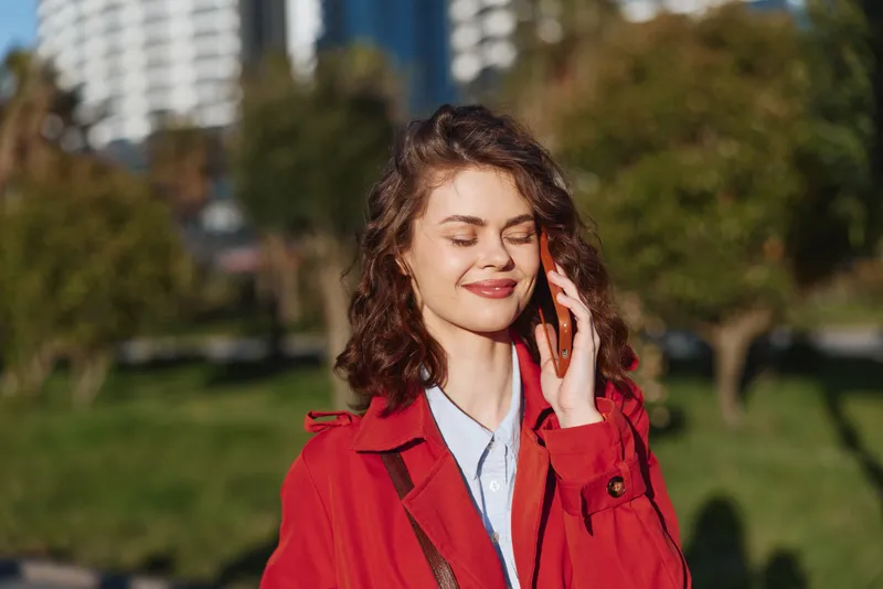 A young woman in a red coat smiles with her eyes closed in a bright city park, enjoying the warm sun while distant buildings rise behind her, creating a relaxed urban portrait.
