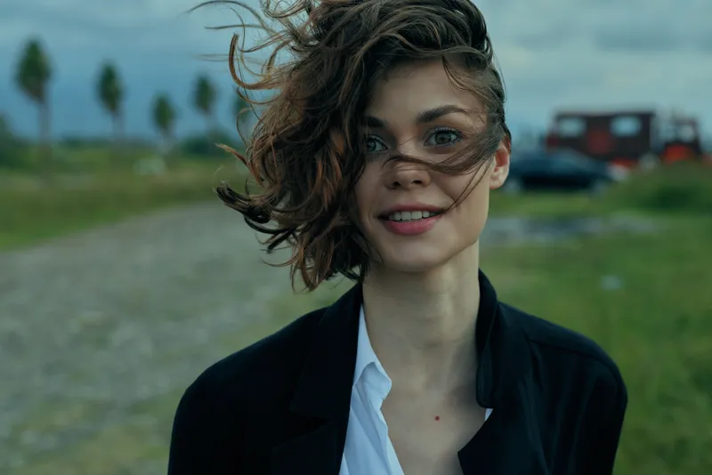Woman in a blazer stands outdoors with windblown hair along a rural road, confident expression, moody sky, casual professional style and open landscape scene.
