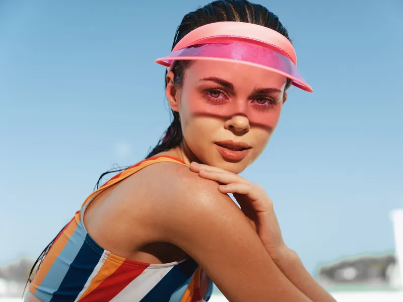 Vibrant portrait of an athletic model in a striped swimsuit and pink visor, sunlit skin and confident gaze, set against a clear blue sky and bright outdoors.