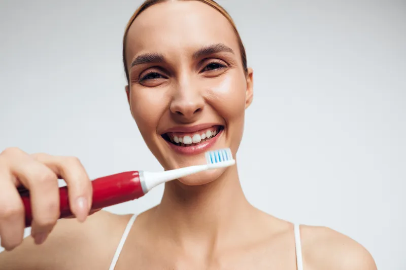 Young woman with brunette hair smiles happily while holding a red electric toothbrush close to her face against a plain white background.
