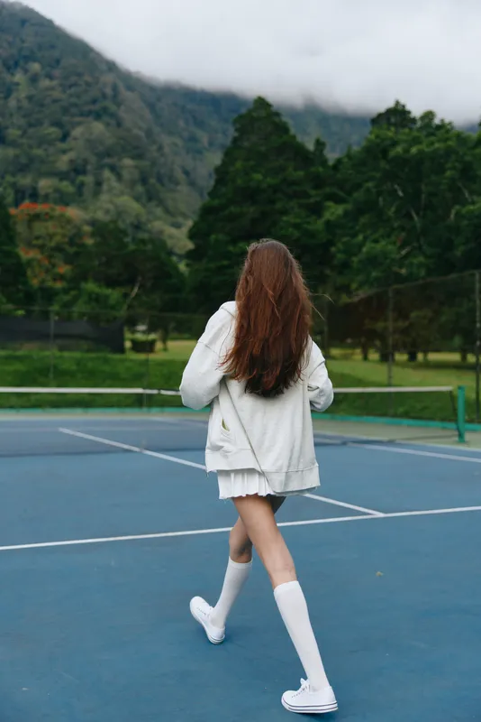 A young woman in white sporty attire walks on a tennis court surrounded by lush greenery, showcasing a healthy and active lifestyle in a serene setting