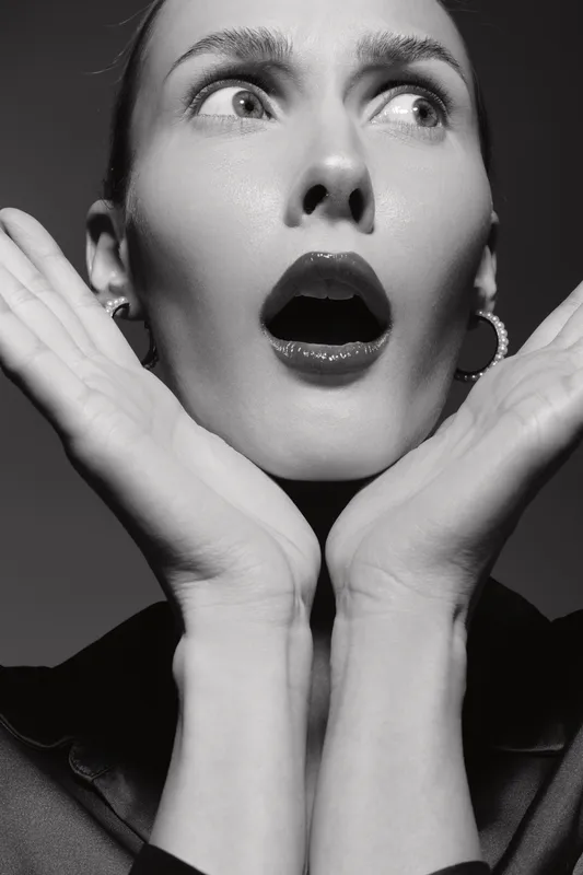 Surprised white woman with pale skin and short hair holds hands near face with open mouth. Black and white portrait with dramatic expression and high contrast lighting.