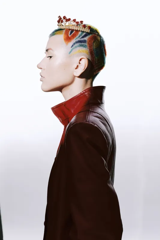 Woman with colorful shaved hair wearing a crown and a red leather jacket, showing bold style and artistic self-expression in a minimalist studio setting.