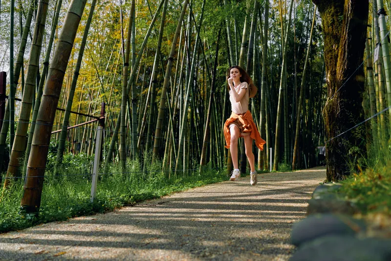 Woman running on a gravel path through a dense bamboo forest, jogging for fitness on a scenic trail outdoors. Young athlete in casual clothing and sneakers enjoying nature and motion.
