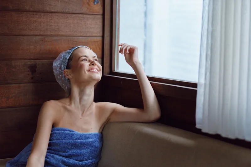 Relaxed woman in towel smiling and enjoying natural light by window inside wooden spa room, cozy atmosphere, wellness and self-care concept.