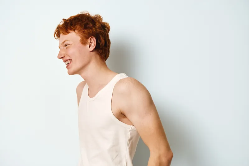 Young man with red hair smiling in tank top on white background, side view of happy male model laughing indoors with casual clothing and natural light.