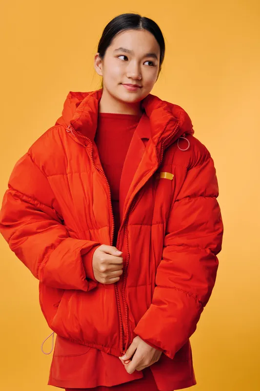 Young woman wearing bright red padded jacket and sweater standing against solid orange background looking sideways with slight smile, casual winter outfit and fashion style portrait.