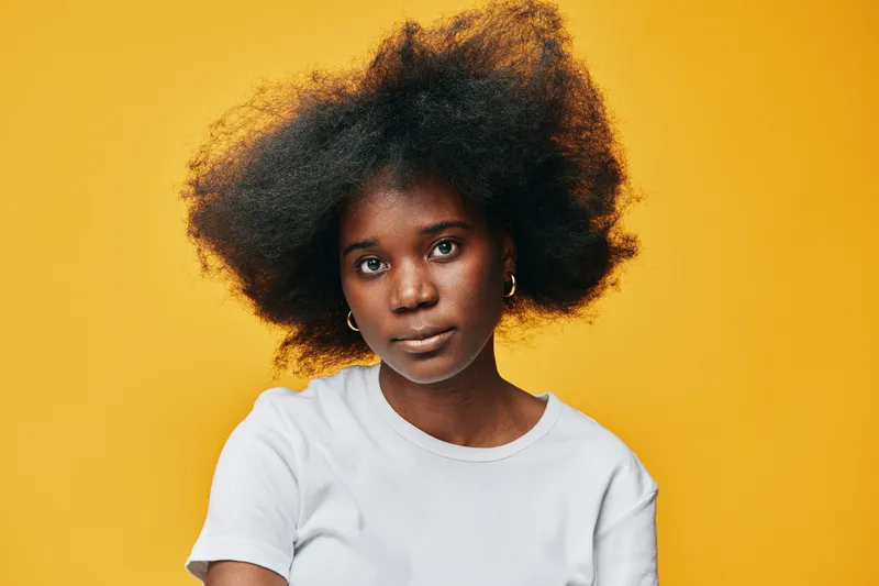 Young Black woman with voluminous natural hair, wearing a white t shirt, expressing confidence against a vibrant yellow background