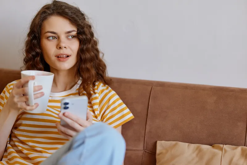 A young woman with curly hair sits on a brown sofa, holding a coffee mug and scrolling through her smartphone in a cozy home setting, suggesting a casual morning routine.