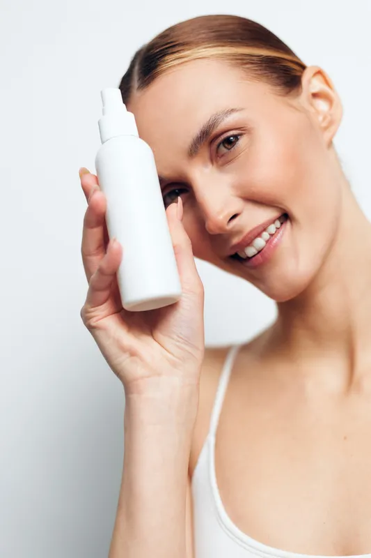 Smiling woman holding white spray bottle near her face on white background. Concept of skincare, beauty, personal care, and wellness with natural light and soft expression.