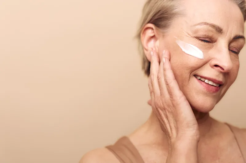 Smiling mature woman applies moisturizing cream on her cheek with closed eyes, enjoying skincare routine. Concept of natural beauty, self-care, and healthy skin at home.