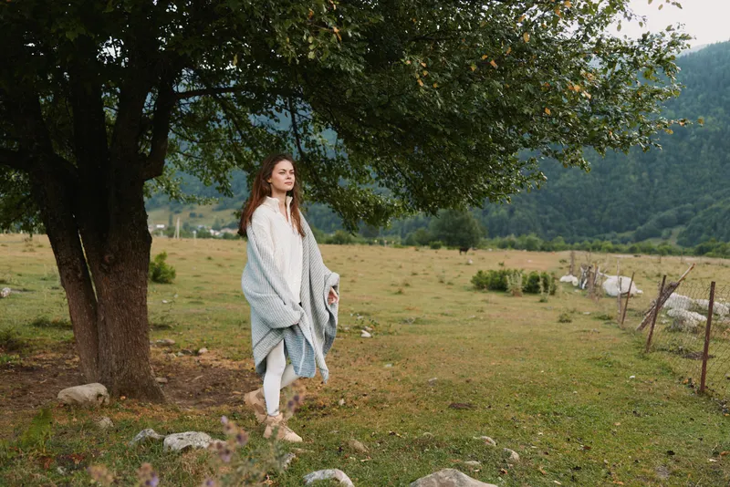 Young woman wrapped in a blanket standing barefoot under a tree in a green meadow with mountains in the background. Concept of lifestyle, nature, and relaxation.