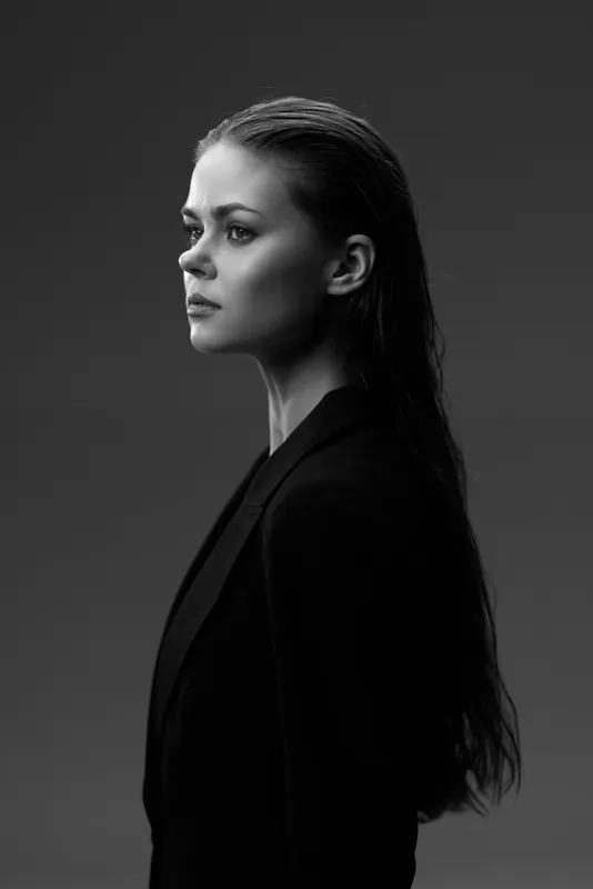 Black and white portrait of a serious woman in a dark blazer with long hair, looking thoughtful and focused in a studio setting.