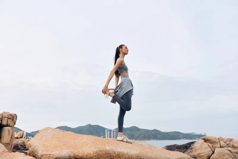 Young Asian woman practicing yoga outdoors on rocky terrain, wearing sportswear, showcasing flexibility and serenity with a serene backdrop of mountains