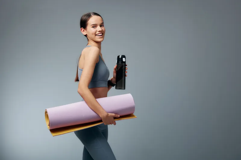 Fitness minded woman in athletic wear carries a yogamat and waterbottle, ready for a workout session, showcasing balance, energy and motivation in a clean studio setting.