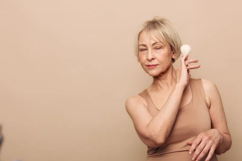 Mature woman with short blonde hair applies soft powder puff to her face on beige background. Concept of skincare, beauty routine, and natural self-care for aging skin.