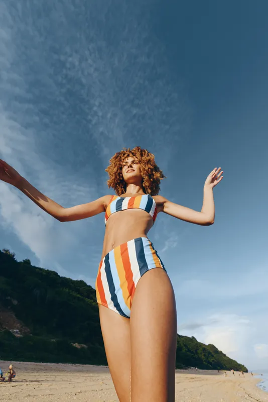 Young woman with curly hair wearing a colorful striped bikini stands on a sandy beach with arms raised under a clear blue sky, enjoying a sunny summer day.