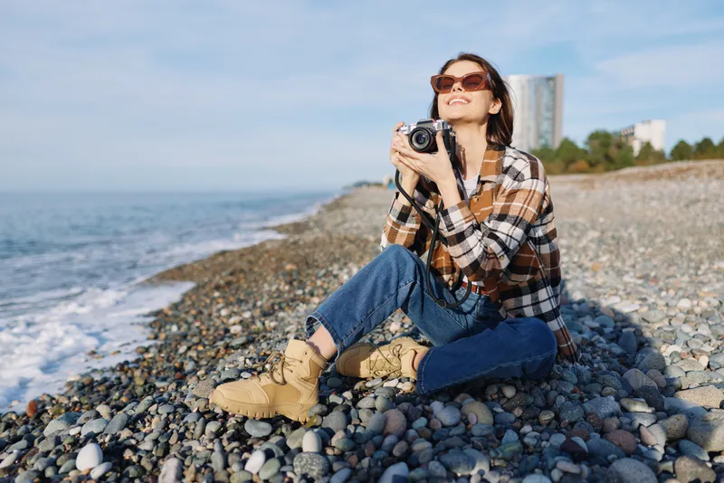 Photographer, camera, beach, rocks, woman, outdoors on a rocky shore wearing casual plaid shirt, jeans and boots under a sunny sky by the sea capturing a creative moment